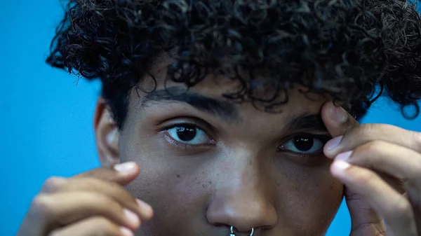 Vue recadrée de l'homme afro-américain touchant les sourcils et regardant la caméra isolée sur bleu — Photo de stock