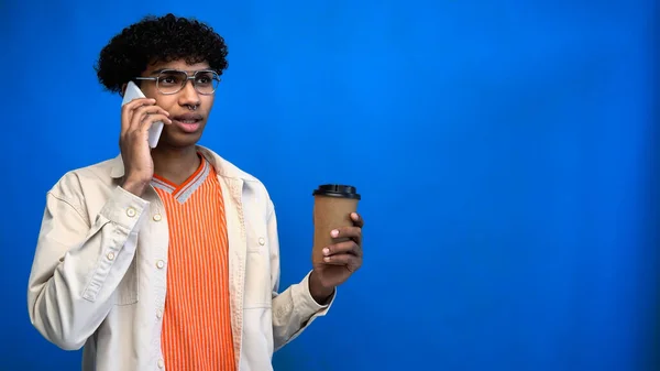 Homem americano africano elegante falando no smartphone e segurando café para ir no fundo azul — Fotografia de Stock
