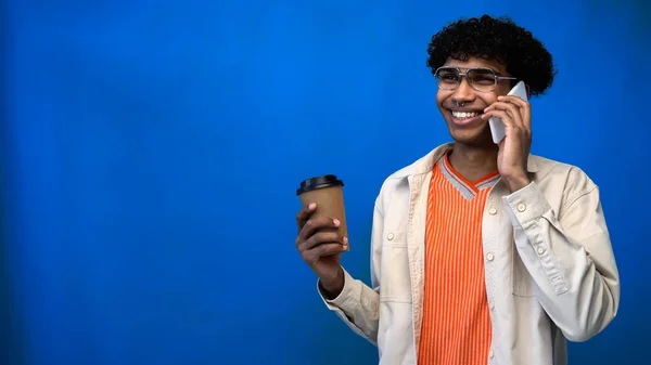 Allegro uomo afroamericano con gli occhiali che parla al cellulare e tiene il caffè per andare su sfondo blu — Foto stock