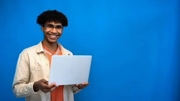 Улыбающийся африканский американский фрилансер смотрит в камеру и держит ноутбук на синем фоне — стоковое фото