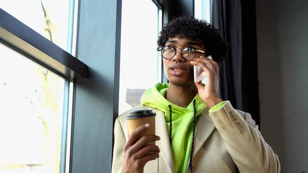 Молодий афроамериканець, який розмовляє по мобільному телефону і тримає каву біля вікна в офісі — стокове фото
