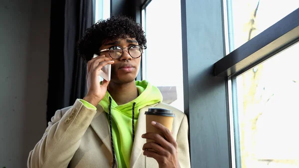 Uomo d'affari afroamericano in occhiali che tiene il caffè per andare a parlare sullo smartphone vicino alla finestra in ufficio — Foto stock