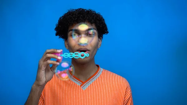Jovem afro-americano soprando bolhas de sabão e olhando para a câmera isolada em azul — Fotografia de Stock