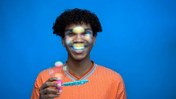 Sorridente uomo africano americano che tiene la bacchetta vicino alle bolle di sapone isolate sul blu — Foto stock