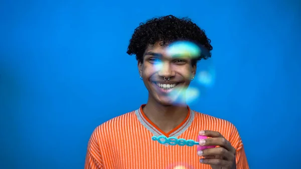 Alegre homem americano africano segurando varinha e olhando para a câmera perto de bolhas de sabão isolado em azul — Fotografia de Stock