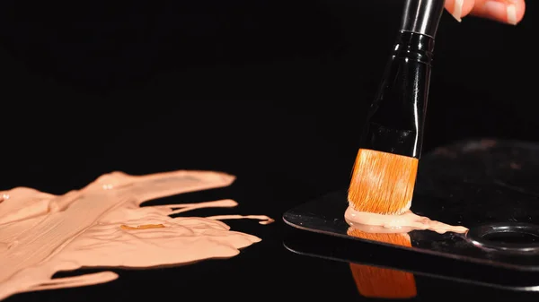 Vue rapprochée de la femme tenant une brosse cosmétique près du fond de teint sur fond noir — Photo de stock