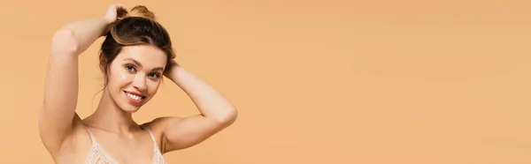 Pretty and happy woman in bra looking at camera and fixing hair isolated on beige, banner — Stock Photo