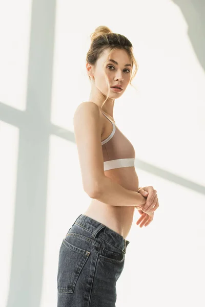 Young and slender woman in sports top looking at camera on white background with shadows — Stock Photo