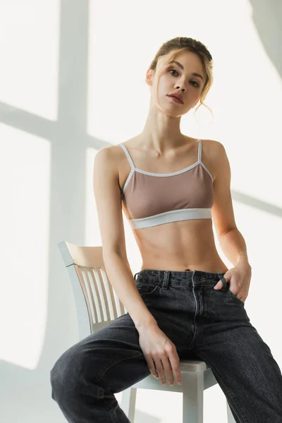 Mujer en top deportivo y jeans sentados en silla con la mano en el bolsillo y mirando a la cámara sobre fondo blanco con sombras - foto de stock