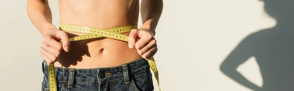 Partial view of slim woman in jeans measuring waist on white background with shadow, banner — Stock Photo