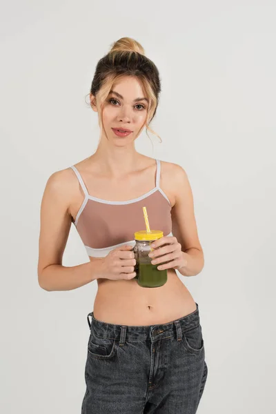 Slender woman in sports top holding fresh smoothie and looking at camera isolated on grey — Stock Photo
