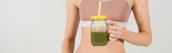 Partial view of fit woman in sports top holding jar of fresh smoothie isolated on grey, banner — Stock Photo