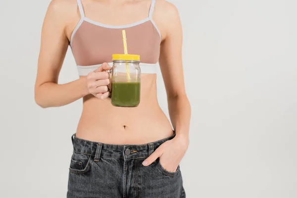 Abgeschnittene Ansicht einer jungen und fitten Frau, die Smoothie hält, während sie mit der Hand in der Tasche auf grau isoliert steht — Stockfoto