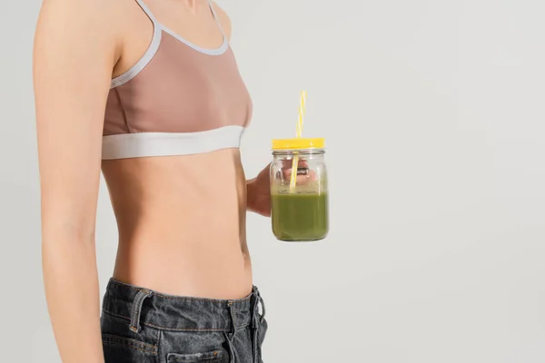 Vista ritagliata della donna in forma in sport top holding frullato fresco isolato su grigio — Foto stock