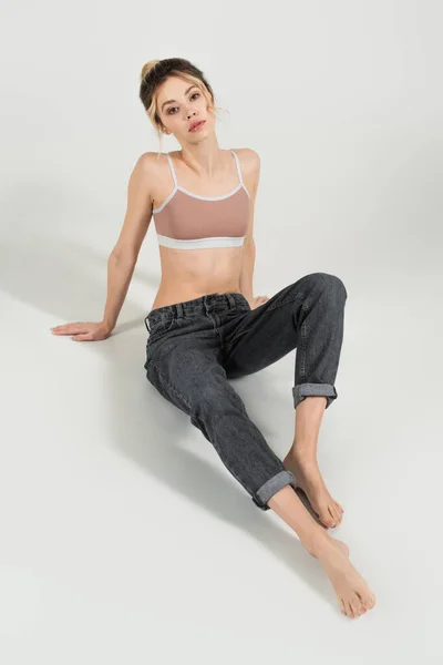 Junge Frau mit perfektem Körper und perfekter Haut sitzt in Jeans und Sporttop auf grauem Hintergrund — Stockfoto