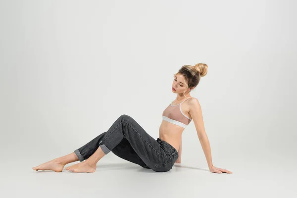 Full length of young barefoot woman with perfect figure sitting in jeans on grey background — Stock Photo