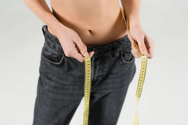 Cropped view of slim woman in jeans measuring waist isolated on grey — Stock Photo