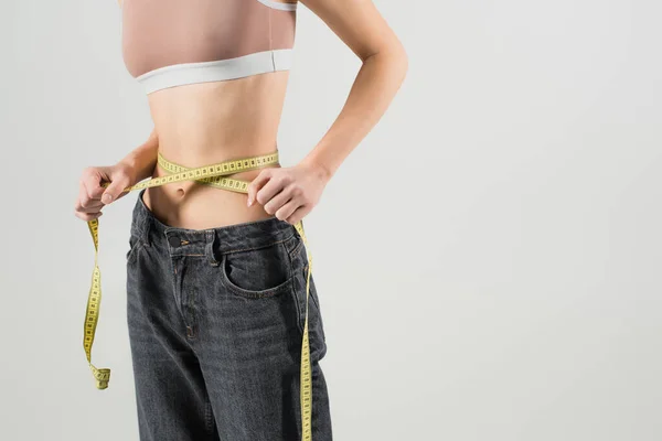 Cropped view of slender woman in sports top and jeans measuring waist isolated on grey — Stock Photo