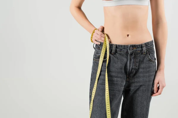Обрізаний вид підходящої жінки в джинсах, що стоїть з рукою на стегні і вимірювальна стрічка ізольована на сірому — стокове фото