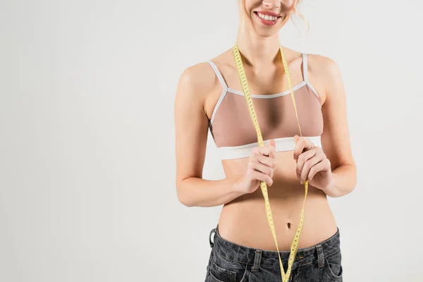 Vue recadrée d'une femme heureuse et en forme dans un haut de sport tenant un ruban à mesurer isolé sur gris — Photo de stock