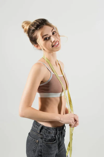 Slender woman in sports top holding measuring tape and smiling at camera isolated on grey — Stock Photo