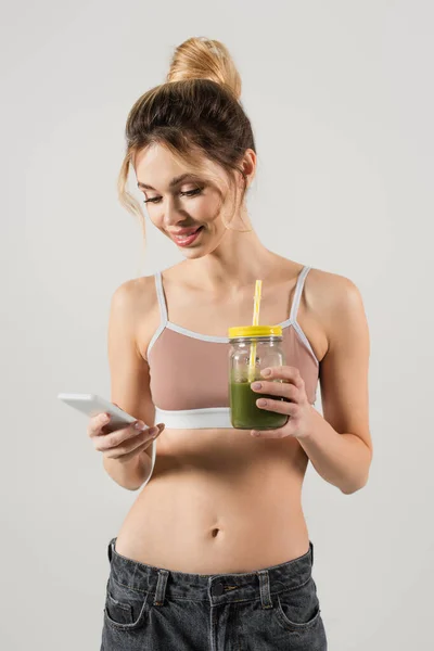Lächelnde Frau mit perfektem Körper und frischem Smoothie mit Smartphone isoliert auf grau — Stockfoto