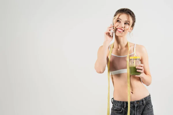 Cheerful woman with measuring tape and jar of smoothie talking on smartphone isolated on grey — Stock Photo