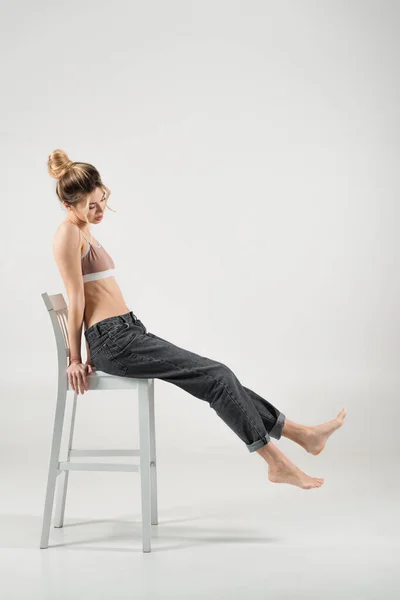 Volle Länge der schlanken barfüßigen Frau in Sport-Top und Jeans sitzt auf Stuhl auf grauem Hintergrund — Stockfoto