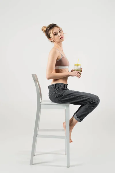 Esbelta mujer en top deportivo y jeans sosteniendo smoothie mientras se sienta en silla blanca sobre fondo gris - foto de stock