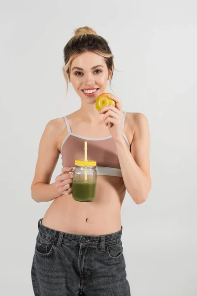 Mujer joven con cara y cuerpo perfectos sosteniendo manzana madura y batido fresco aislado en gris - foto de stock