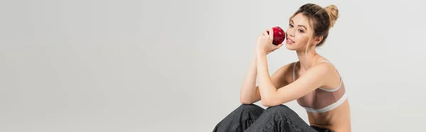 Положительная молодая женщина в спортивной верхней сидя с спелым яблоком изолированы на сером, баннер — стоковое фото
