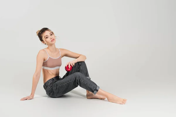 Full length of slender barefoot woman holding juicy apple and sitting on grey background — Stock Photo