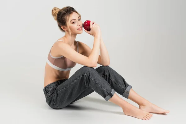 Mujer joven descalza con el cuerpo perfecto y la piel sentada con manzana fresca sobre fondo gris - foto de stock