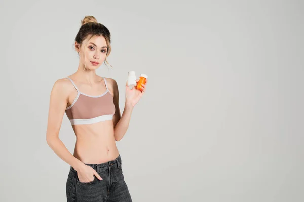 Fit woman in sports bra holding containers with vitamins while standing with hand in pocket isolated on grey — Stock Photo