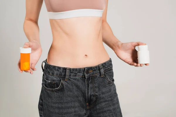 Teilbild einer schlanken Frau in Jeans, die Gefäße mit Vitaminen auf grau isoliert hält — Stockfoto