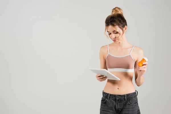 Mujer joven en top deportivo sosteniendo vitaminas y mirando tableta digital aislada en gris - foto de stock