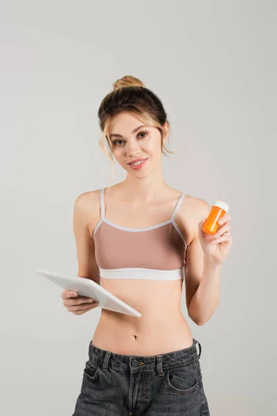 Здоровой женщины в спортивном бюстгальтере с цифровыми таблетками и витаминами, изолированными на сером — стоковое фото