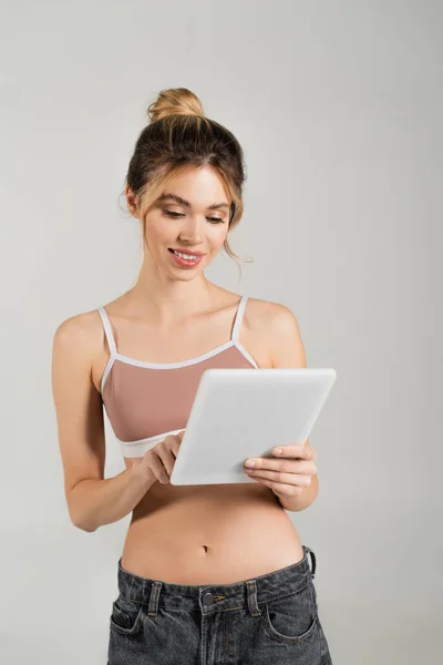 Улыбающаяся женщина с идеальной кожей и стройным телом с помощью цифрового планшета, изолированного на сером — стоковое фото