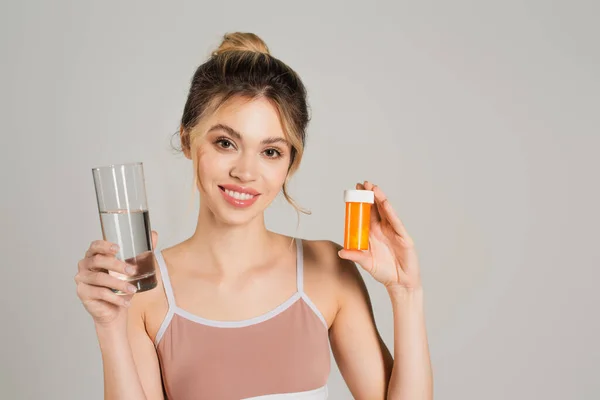 Жінка з ідеальною шкірою посміхається на камеру, тримаючи склянку води і вітаміни ізольовані на сірому — стокове фото