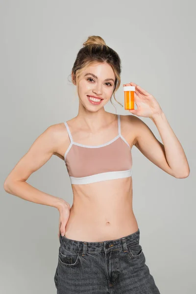 Cheerful and fit woman in sports top standing with hand on hip and showing container with vitamins isolated on grey — Stock Photo