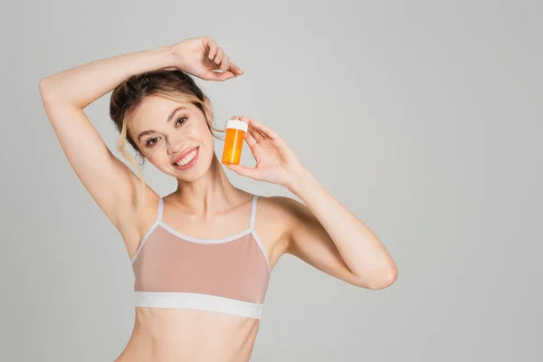 Excited and slender woman with natural makeup holding vitamins and smiling at camera isolated on grey — Stock Photo