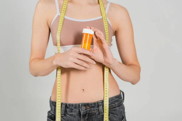 Частичный вид тонкой женщины с измерительной лентой и контейнером с витаминами, изолированными на сером — стоковое фото
