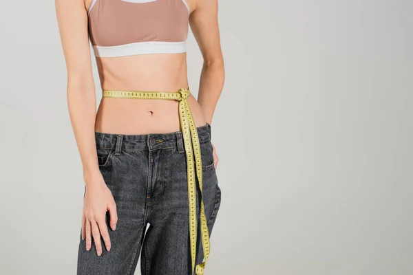 Partial view of fit woman in sports top and jeans standing with measuring tape on waist isolated on grey — Stock Photo