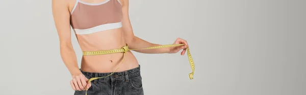 Partial view of sllim woman in sports top with measuring tape on waist isolated on grey, banner — Stock Photo