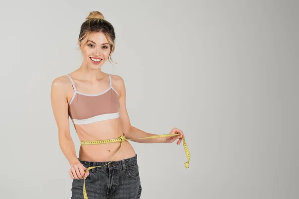 Pleased and slender woman with measuring tape on waist smiling at camera isolated on grey — Stock Photo