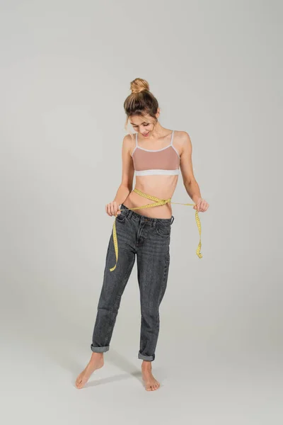 Full length of barefoot woman in jeans and sports top measuring waist on grey background — Stock Photo