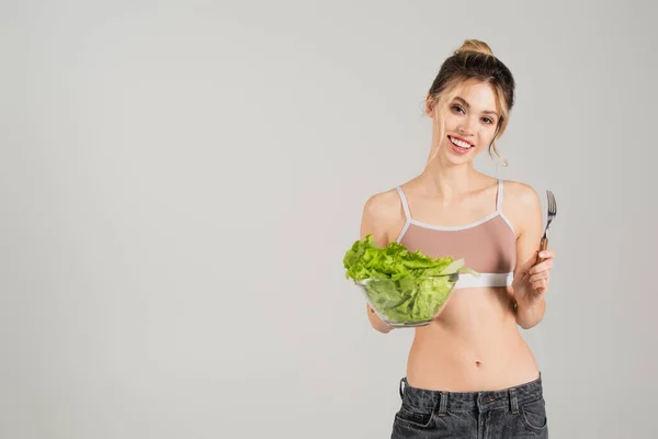 Молодая женщина с стройным телом, держа вилку и свежий салат, улыбаясь в камеру, изолированную на сером — стоковое фото
