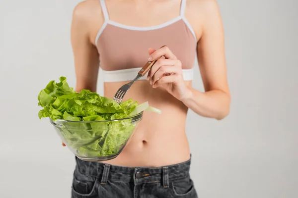 Cropped view of woman in sports top holding fork and fresh lettuce isolated on grey — Stock Photo