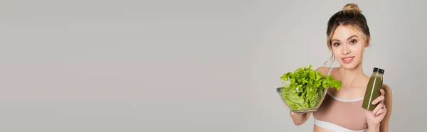 Mulher bonita com tigela de alface e garrafa de smoothie sorrindo para a câmera isolada em cinza, banner — Fotografia de Stock