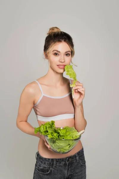 Smiling woman with slim body holding lettuce and looking at camera isolated on grey — Stock Photo